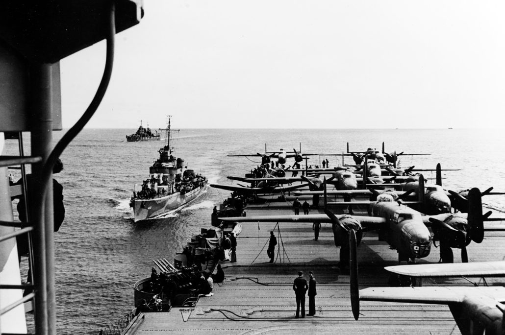 View looking aft from the island of USS Hornet (CV-8), while en route to the mission's launching point. USS Gwin (DD-433) is coming alongside, as USS Nashville (CL-43) steams in the distance. Eight of the mission's sixteen B-25B bombers are parked within view, as are two of the ship's SBD scout bombers. Note midships elevator, torpedo elevator, arresting gear and flight deck barriers in the lower portion of the photo, and 1.1 quad anti-aircraft machine gun mount at left. U.S. Naval History and Heritage Command Photograph.